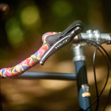 Load image into Gallery viewer, Close-up of a bicycle handlebar with colorful grips and a Shimano shifter against a blurred natural background.