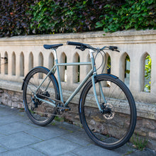 Load image into Gallery viewer, Bicycle leaning against a stone wall with greenery in the background
